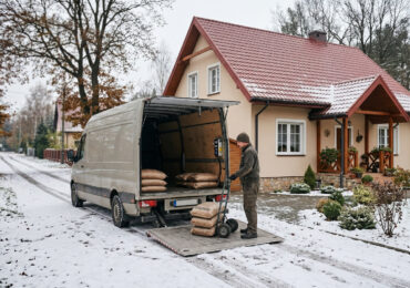 Dostawca rozładowuje worki pelletu z beżowego vana na windę za pomocą wózka ręcznego na ośnieżonej drodze przed budynkiem mieszkalnym.